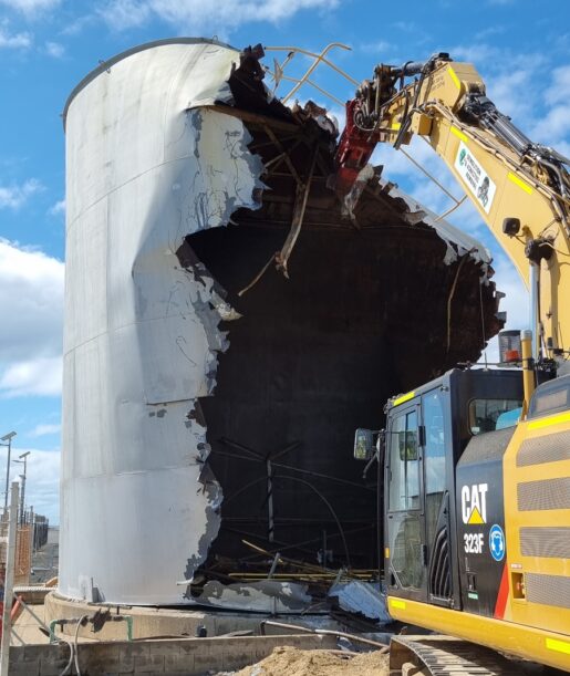 CAT 323F excavator demolishing a large steel storage tank during an industrial demolition project by Razor Asbestos.