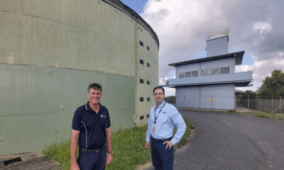 Two workers standing in front of a large green water reservoir tank before demolition.