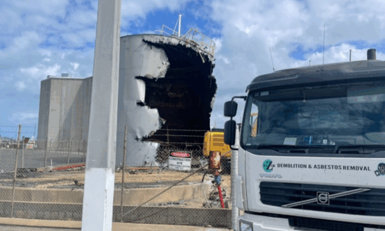 Partially dismantled steel tallow tank with demolition truck positioned nearby.