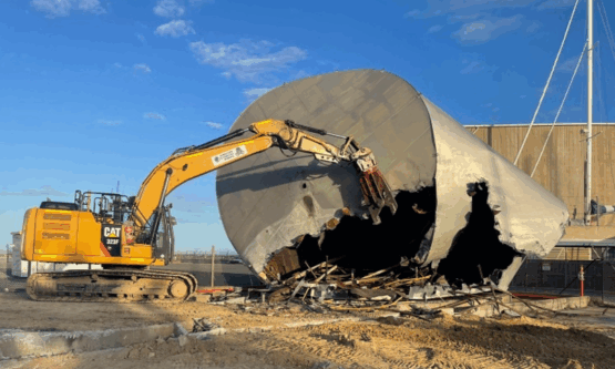 Excavator lifting a large circular steel tank segment during demolition operations.